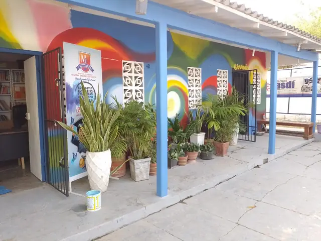 Corredor del colegio con columnas azules, un mural de colores y materas con plantas.