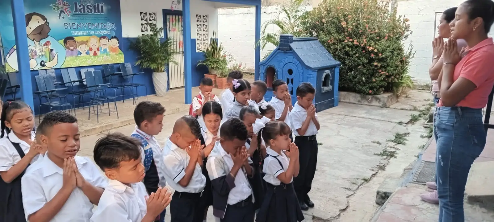 Estudiantes de primaria en uniforme oran con las manos juntas en el patio del colegio, acompañados por dos docentes.